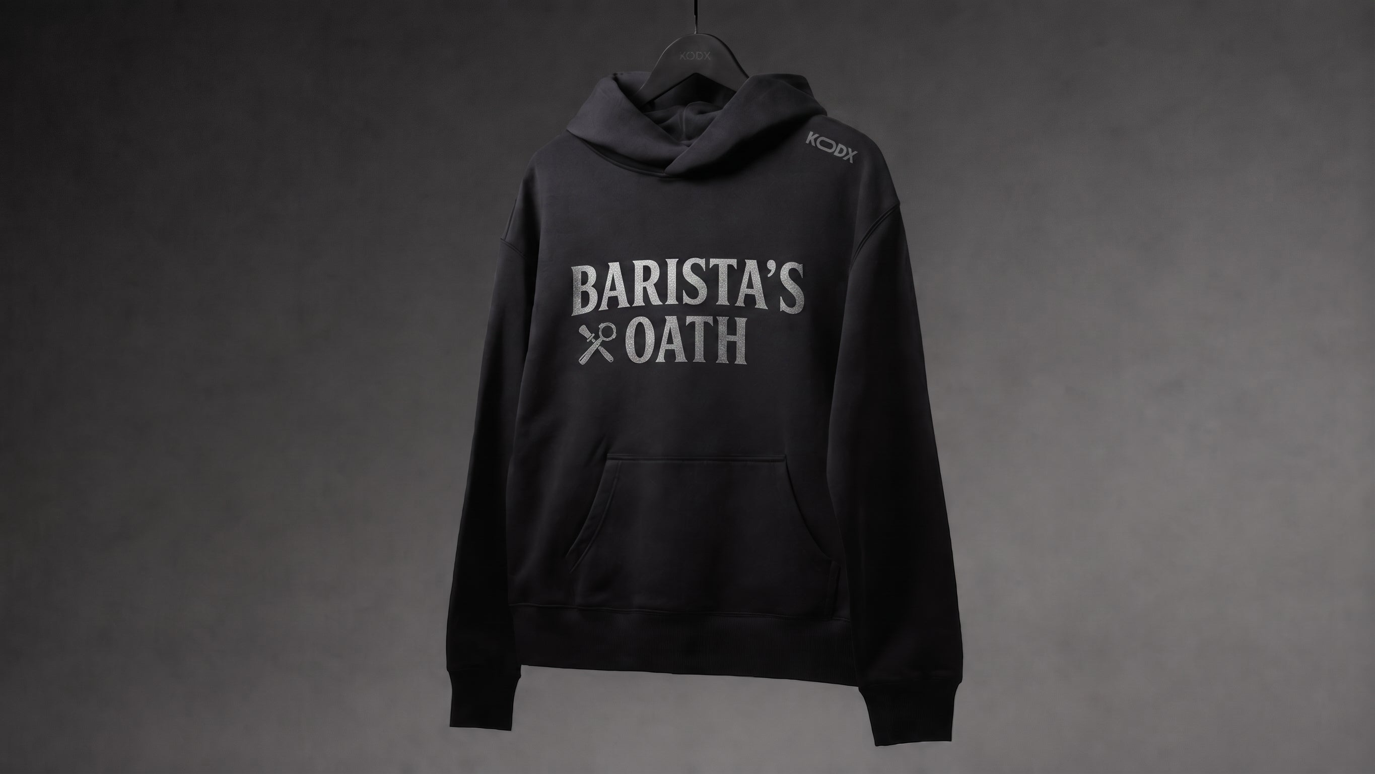 A black pullover hoodie hanging on a black hanger against a dark gray textured background. The hoodie features 'BARISTA'S OATH' printed in white across the chest, with a small white logo on the left shoulder.